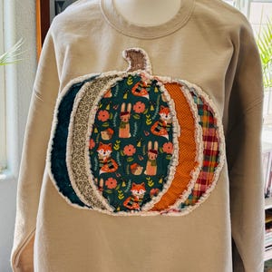 May include: A beige crewneck sweatshirt featuring a large, central pumpkin applique. The pumpkin is constructed from various fabric segments, including a dark green print with foxes and rabbits, a mustard orange polka dot, and a red plaid pattern.