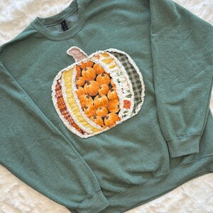 May include: A heathered sage green crewneck sweatshirt featuring a large, central pumpkin-shaped appliqu&eacute;. The pumpkin is constructed from various fabric strips in orange, yellow, white, and dark green plaid, with frayed edges.
