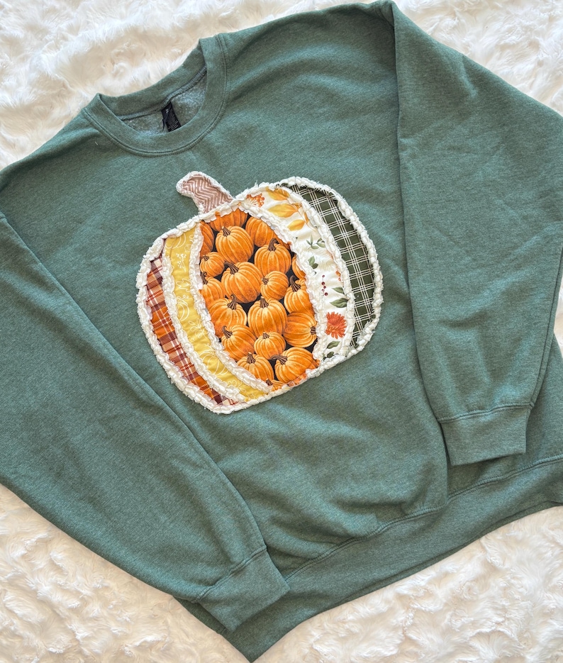 May include: A heathered sage green crewneck sweatshirt featuring a large, central pumpkin-shaped appliqu&eacute;. The pumpkin is constructed from various fabric strips in orange, yellow, white, and dark green plaid, with frayed edges.