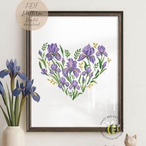 Puede incluir: Arte de punto de cruz enmarcado que representa un corazón compuesto por flores de iris moradas y hojas verdes. La obra de arte está en un marco marrón oscuro. El texto "PDF Pattern Digital download" está en la esquina superior izquierda.