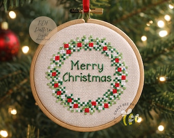 May include: A festive cross-stitch Christmas ornament. It features a wreath design in red and green, with the words "Merry Christmas" in green. The ornament is in a wooden hoop and suspended by a red ribbon.