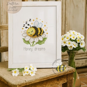 May include: A framed cross-stitch art piece featuring a sleeping bee on a flower, with the words "Honey dreams" below. The bee is yellow and black, with blue wings. The frame is white, and the art is displayed on a wooden chair.