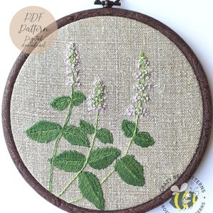 Könnte beinhalten: Gesticktes Blumendesign in einem runden Holzreifen. Das Kunstwerk zeigt grüne Blätter und Stiele mit zarten lila Blütenähren. Der Text "PDF Pattern Digital download" befindet sich in einem Kreis oben links.
