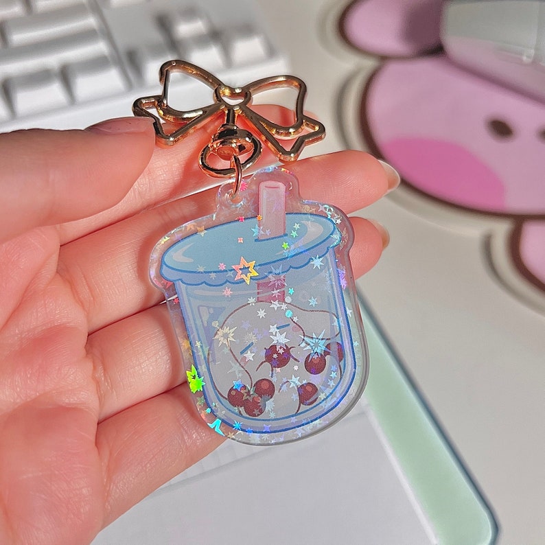 Puede incluir: Un llavero transparente e iridiscente con forma de vaso de boba con una pajita y perlas de boba en su interior. El llavero tiene un lazo dorado sujeto a la parte superior.