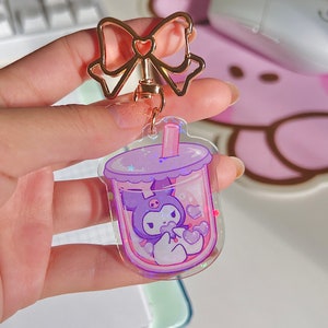 Puede incluir: Un llavero de acr&iacute;lico transparente morado y rosa con un personaje de dibujos animados dentro de una taza de t&eacute; de burbujas. El personaje es un gato blanco y negro con un lazo rosa y un coraz&oacute;n rosa en su pata. El llavero tiene un lazo dorado y una cadena dorada.