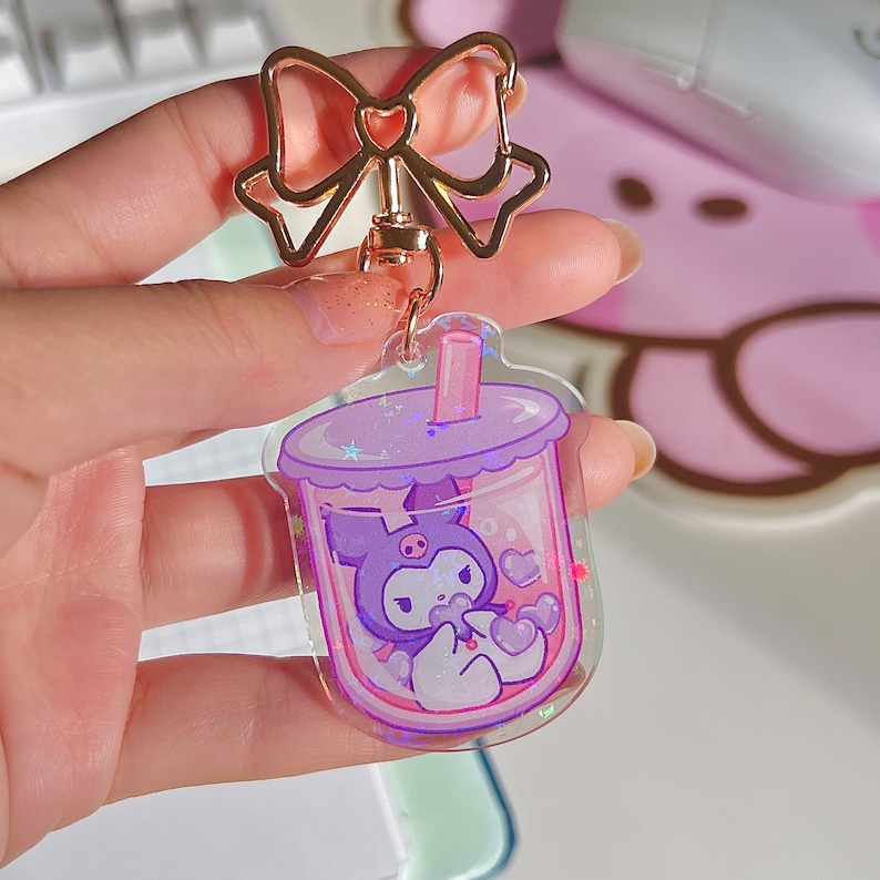 Puede incluir: Un llavero de acr&iacute;lico transparente morado y rosa con un personaje de dibujos animados dentro de una taza de t&eacute; de burbujas. El personaje es un gato blanco y negro con un lazo rosa y un coraz&oacute;n rosa en su pata. El llavero tiene un lazo dorado y una cadena dorada.