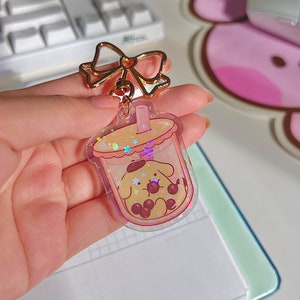 Puede incluir: Un llavero transparente con un perro de dibujos animados dentro de una taza de t&eacute; de burbujas. El llavero tiene un lazo dorado y una cadena dorada. El fondo es una superficie rosa y blanca.