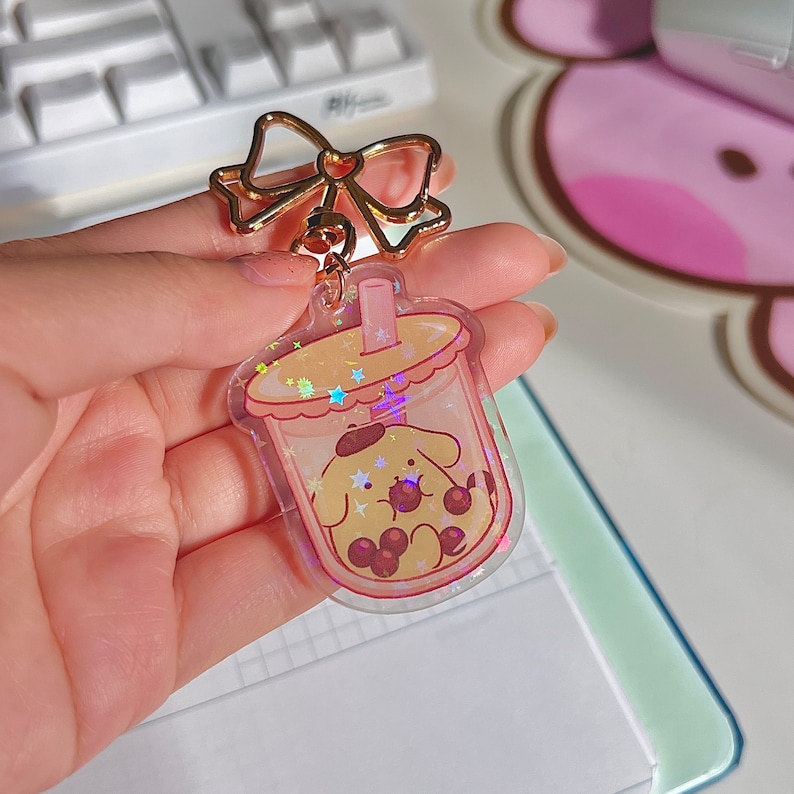 Puede incluir: Un llavero transparente con un perro de dibujos animados dentro de una taza de t&eacute; de burbujas. El llavero tiene un lazo dorado y una cadena dorada. El fondo es una superficie rosa y blanca.