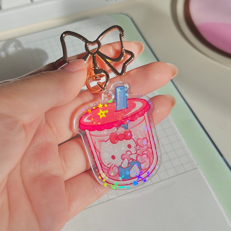 Puede incluir: Un amuleto iridiscente rosa y rojo con un personaje de Hello Kitty dentro de una taza de boba. El amuleto tiene un lazo dorado y una cadena dorada.