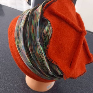 Peut inclure: Un chapeau orange brûlé avec un bandeau rayé coloré. Le chapeau a une couronne arrondie et un bord plié avec des coutures noires. Le bandeau présente des rayures verticales dans les tons de vert, de bleu et de jaune.