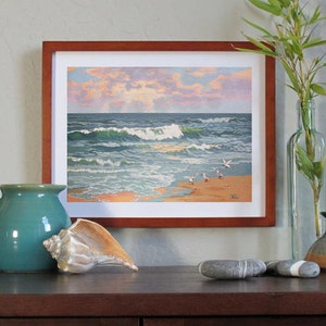 May include: Framed artwork depicting a coastal scene with crashing waves, seagulls, and a sunset. A teal ceramic pitcher, a seashell, and decorative stones are in the foreground. A clear glass bottle with bamboo and a small ceramic vase are also present.
