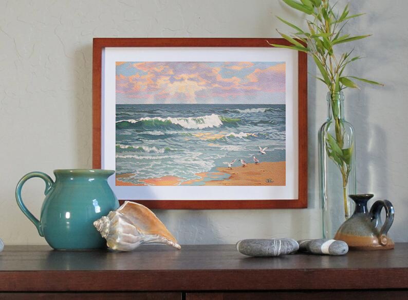 May include: Framed artwork depicting a coastal scene with crashing waves, seagulls, and a sunset. A teal ceramic pitcher, a seashell, and decorative stones are in the foreground. A clear glass bottle with bamboo and a small ceramic vase are also present.