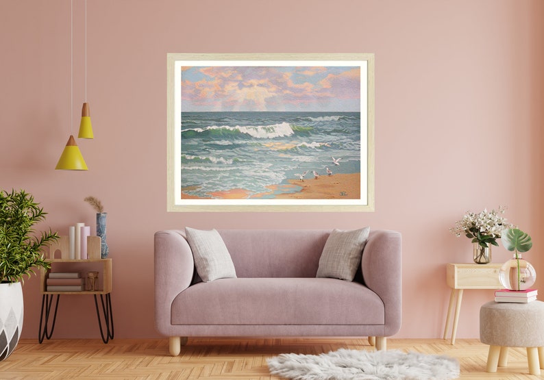May include: Framed artwork depicting a coastal scene with ocean waves, seagulls, and a sandy beach. The painting features a light-colored wooden frame and is displayed above a pink sofa. The artwork captures a serene beach landscape.