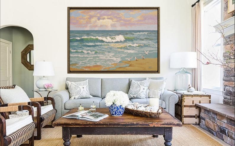 May include: A living room scene featuring a framed seascape painting above a gray sofa with decorative pillows. A dark wood coffee table holds a blue and white vase with white flowers. Two wicker chairs and a side table with a lamp complete the decor.
