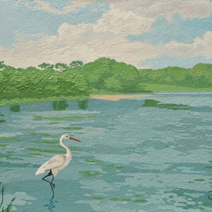May include: A white egret stands in a blue lake with green trees in the background. The sky is blue with white clouds.