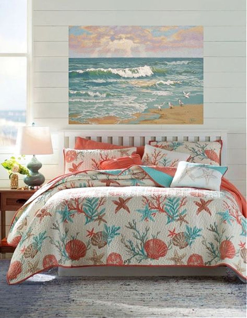 May include: A bed with a white headboard and a coastal-themed quilt. The quilt features a pattern of coral, seashells, and starfish in shades of coral, teal, and beige. A painting of a beach scene hangs above the bed.