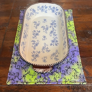 May include: An oval ceramic baking dish with a white interior and blue floral patterns, featuring brown handles with white floral accents. The dish rests on a rectangular fabric mat with a green and purple gradient background and black spider web print.