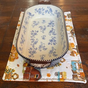 May include: An oval ceramic baking dish with a cream-colored interior featuring a blue floral pattern and a dark brown exterior with a beaded rim. The dish has two handles and sits on a rectangular quilted fabric mat with a fall-themed harvest print.