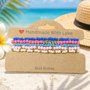Op de afbeelding: Een set kralenarmbanden in blauw, roze en wit, met gouden accenten. De armbanden worden gepresenteerd op een bruine kaart met de tekst "Handmade With Love" en "Best Wishes". De achtergrond toont een strandscène en een strohoed.