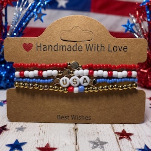 Könnte beinhalten: Ein Set aus vier Armbändern mit roten, weißen, blauen und goldenen Perlen, präsentiert auf einer braunen Karte mit den Texten "Handmade With Love" und "Best Wishes". Die Armbänder bilden mit Buchstabenperlen das Wort "USA". Der Hintergrund zeigt eine amerikanische Flagge und sternförmige Dekorationen.