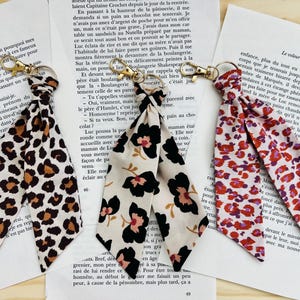 Può includere: Tre portachiavi in tessuto con chiusure color oro sono appoggiati su pagine di testo. I portachiavi presentano diversi motivi: una stampa leopardata marrone e beige, uno sfondo crema con un design floreale nero e rosa e un motivo floreale rosso e rosa.