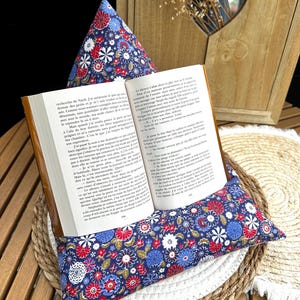 Può includere: Un leggio blu navy con un motivo floreale rosso, bianco e blu tiene un libro aperto. Il leggio è su un tappetino intrecciato, con un mobile in legno e fiori secchi sullo sfondo. Le pagine del libro sono visibili.