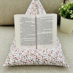 Può includere: Un leggio in tessuto a forma di piramide con sfondo bianco e un motivo floreale in scala ridotta nei toni del marrone, rosa e verde. Un libro tascabile aperto con pagine bianche e testo stampato in nero poggia sulla parte anteriore.