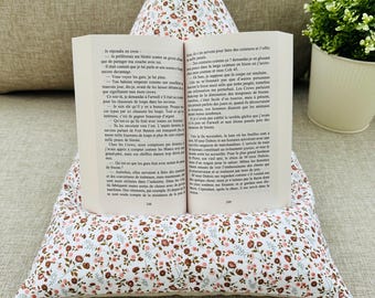 Peut inclure: Un support de livre en tissu en forme de pyramide avec un fond blanc et un motif floral à petite échelle dans des tons marron, rose et vert. Un livre de poche ouvert avec des pages blanches et du texte noir imprimé repose sur le support.