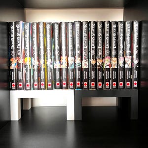 Puede incluir: Una colección de volúmenes de manga "Demon Slayer" expuestos en una estantería. Las lomos son negros con letras blancas e ilustraciones coloridas. Los libros están colocados en una estantería blanca y negra.