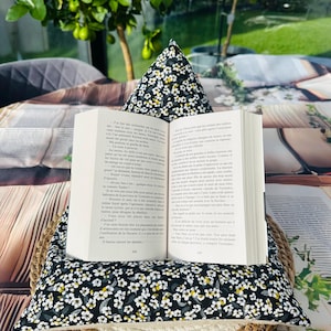 Può includere: Un cuscino per libri con un motivo floreale in bianco e nero, con un libro aperto sopra. Il cuscino è su un cestino intrecciato. Il libro mostra del testo. Un piccolo albero è visibile sullo sfondo.