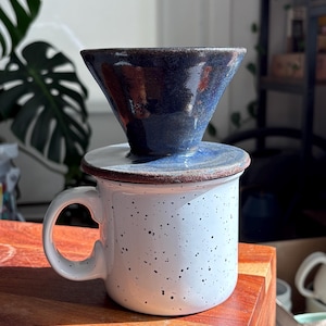 May include: A ceramic pour-over coffee maker with a dark blue cone-shaped filter sits atop a white speckled mug. The mug has a large handle and a brown rim. The coffee maker and mug are on a wooden surface.