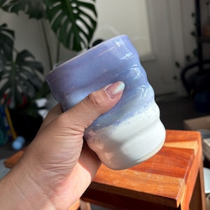 May include: A handmade ceramic mug with a wavy design. The mug features a gradient of colors, transitioning from a light blue at the top to white at the bottom. The mug is held in a person's hand.