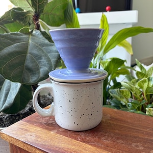 May include: A ceramic coffee pour-over dripper in a light periwinkle blue color with a ribbed texture, resting on top of a white ceramic mug featuring a black speckled pattern and a curved handle, designed for brewing coffee directly into the cup.