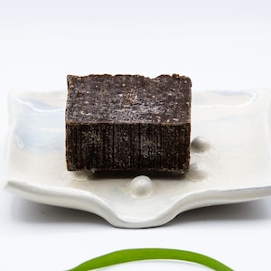 Peut inclure: Un pain de savon rectangulaire brun foncé repose sur un porte-savon carré blanc aux bords ondulés. Le porte-savon comporte trois petits accents ronds blancs. Le savon a une surface texturée.