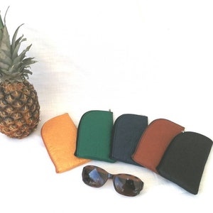 Könnte beinhalten: Eine Auswahl an farbenfrohen Brillenetuis in Orange, Grün, Marineblau, Braun und Schwarz, mit einer Sonnenbrille. Eine Ananas befindet sich links von den Etuis auf einer weißen Oberfläche.