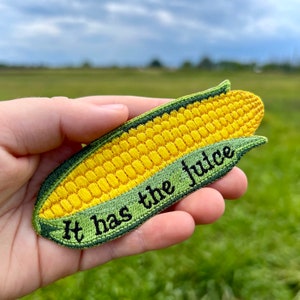 May include: A yellow and green embroidered patch shaped like an ear of corn with the text "It has the juice" on it.