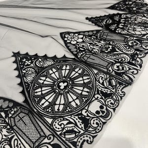 Puede incluir: Tela gótica en blanco y negro con intrincados detalles de encaje. El diseño presenta un patrón repetitivo de vidrieras, ataúdes, calaveras y elementos florales. La tela es transparente, con una textura delicada y aireada.