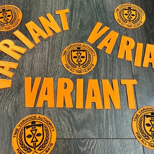 May include: Orange fabric patches with the word "VARIANT" in large letters. The patches are on a wood surface. There are also smaller circular patches with the text "TIME VARIANCE AUTHORITY" and a black and white logo.