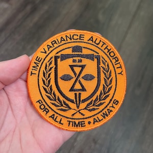 May include: Orange embroidered patch with black text that reads "Time Variance Authority" and "For All Time Always". The patch features a black shield with a stylized hourglass and dagger.