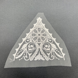 May include: White embroidered triangular patch featuring a gothic design. The patch includes a central circular element, two skull motifs, and ornate scrollwork. The patch is made of a sheer, lightweight fabric.