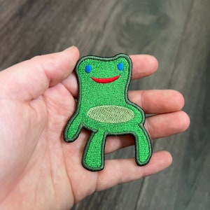 May include: Green felt frog chair patch with blue eyes and a red smile. The frog is sitting on its hind legs with its front legs outstretched.