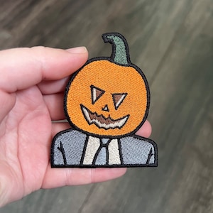 May include: A felt patch of a pumpkin wearing a suit. The pumpkin is orange with a black stitched-on face. The suit is gray with a white collar and tie.