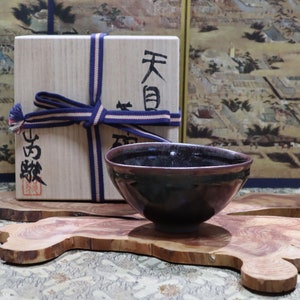 May include: A dark brown tea bowl with a black glaze sits on a wooden stand. The bowl is tied with a blue and white ribbon and placed in front of a wooden box with Japanese characters that read "天目" and "古美術".