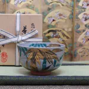 May include: A blue and gold ceramic tea bowl with a bamboo design, sitting on a green mat with a brown box tied with a white ribbon in the background.