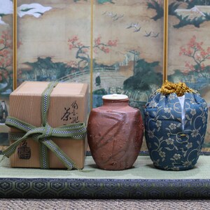 Op de afbeelding: Een bruine doos met een groen lint en Japanse karakters, een bruine keramische pot met een deksel en een blauwe stoffen tas met een bloemenpatroon, allemaal op een groene mat.