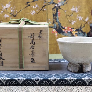 May include: A Japanese tea set comprising a pale ceramic bowl and a wooden box with Japanese calligraphy. The box is tied with a green ribbon. The set is displayed on a blue and white patterned cushion.