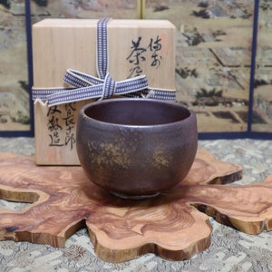 May include: A dark brown ceramic tea bowl with a textured surface sits on a wooden stand. The bowl is in front of a wooden box with a blue and white ribbon and Japanese characters.