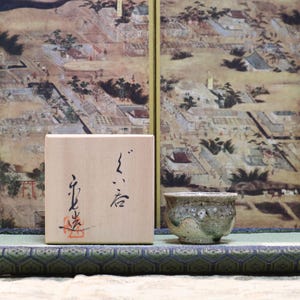 May include: A wooden box with Japanese calligraphy and a small ceramic bowl with a textured surface. The box and bowl are on a green and blue patterned mat, with a painted backdrop.