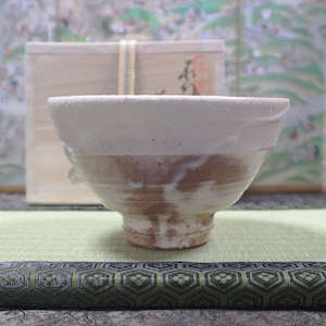 May include: A white ceramic bowl with a brown glaze on the inside. The bowl is sitting on a green mat with a black and white patterned border.