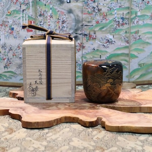 May include: A wooden box with Japanese text and a decorative container with a landscape design, both on a natural wood slab. The box is tied with a blue ribbon. The container is brown and black.